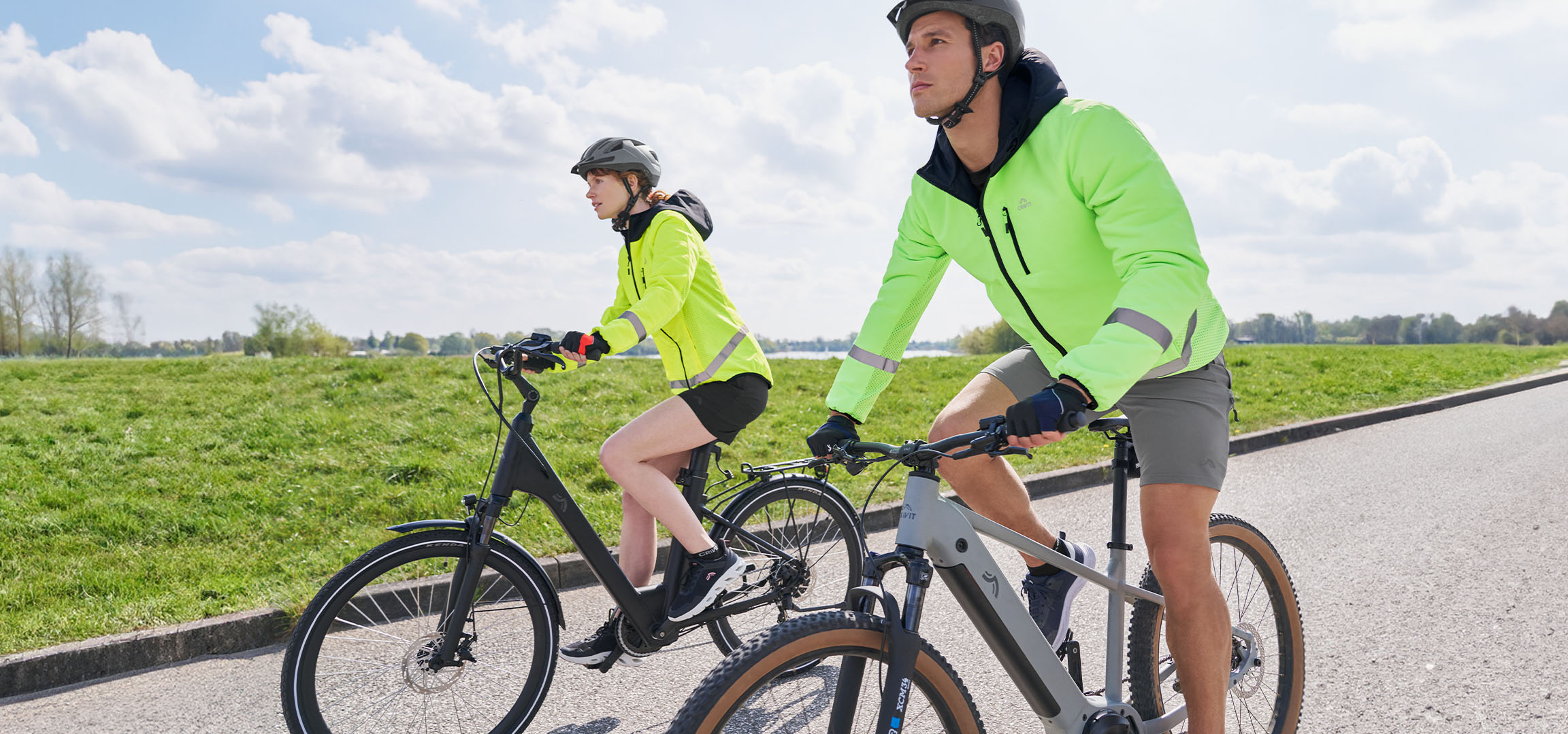 Zwei Radfahrer in leuchtend gelben Jacken fahren auf einem asphaltierten Weg durch eine grüne Landschaft.