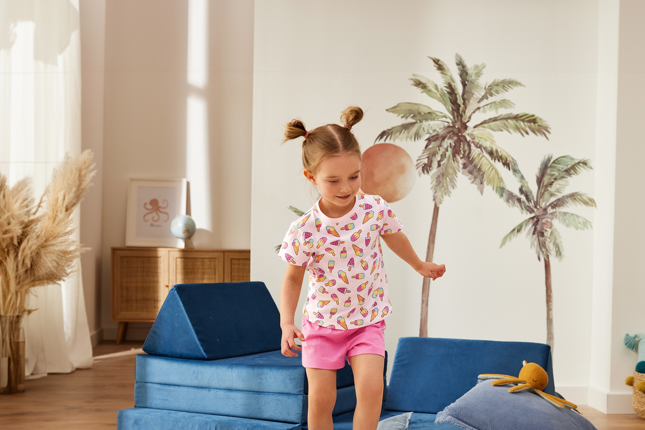 Mädchen in Eiscreme-T-Shirt und Shorts spielt auf blauen Schaumstoffblöcken in einem hellen Raum mit Palmenwandbild.