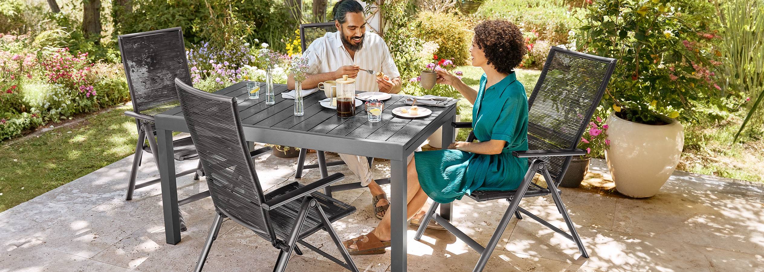 Ein Paar frühstückt an einem grauen Gartentisch mit passenden Stühlen auf einer Terrasse.