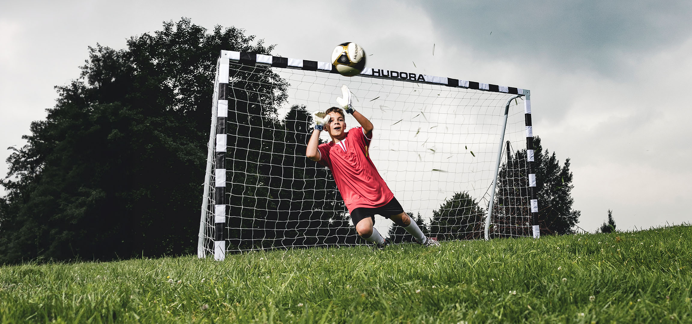 Ein Junge im Torwarttrikot fängt einen Fußball, ein Mädchen springt auf einem Hudora Trampolin.