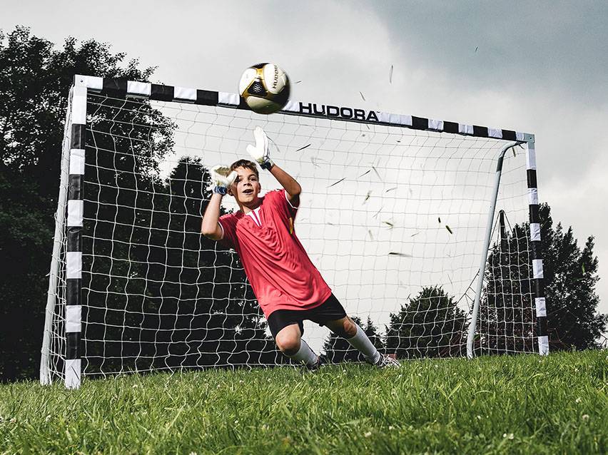 Junge im Torwarttrikot fängt einen Fußball vor einem Hudora-Fußballtor.