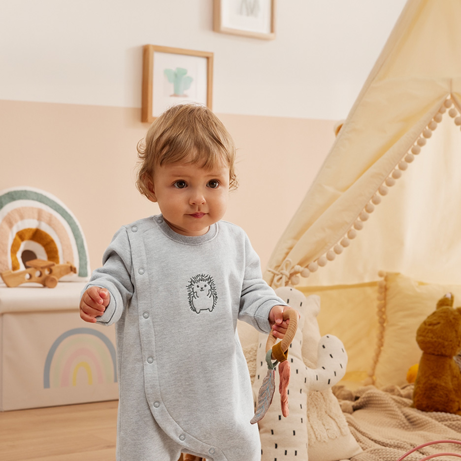 Baby in grauem Strampler mit Igel-Motiv, hält einen Holzbeißring in einem Kinderzimmer.