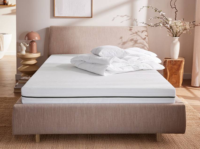 Ein Bett mit einer weißen Matratze, Kissen und Decke in einem Schlafzimmer mit Nachttisch und Vase.