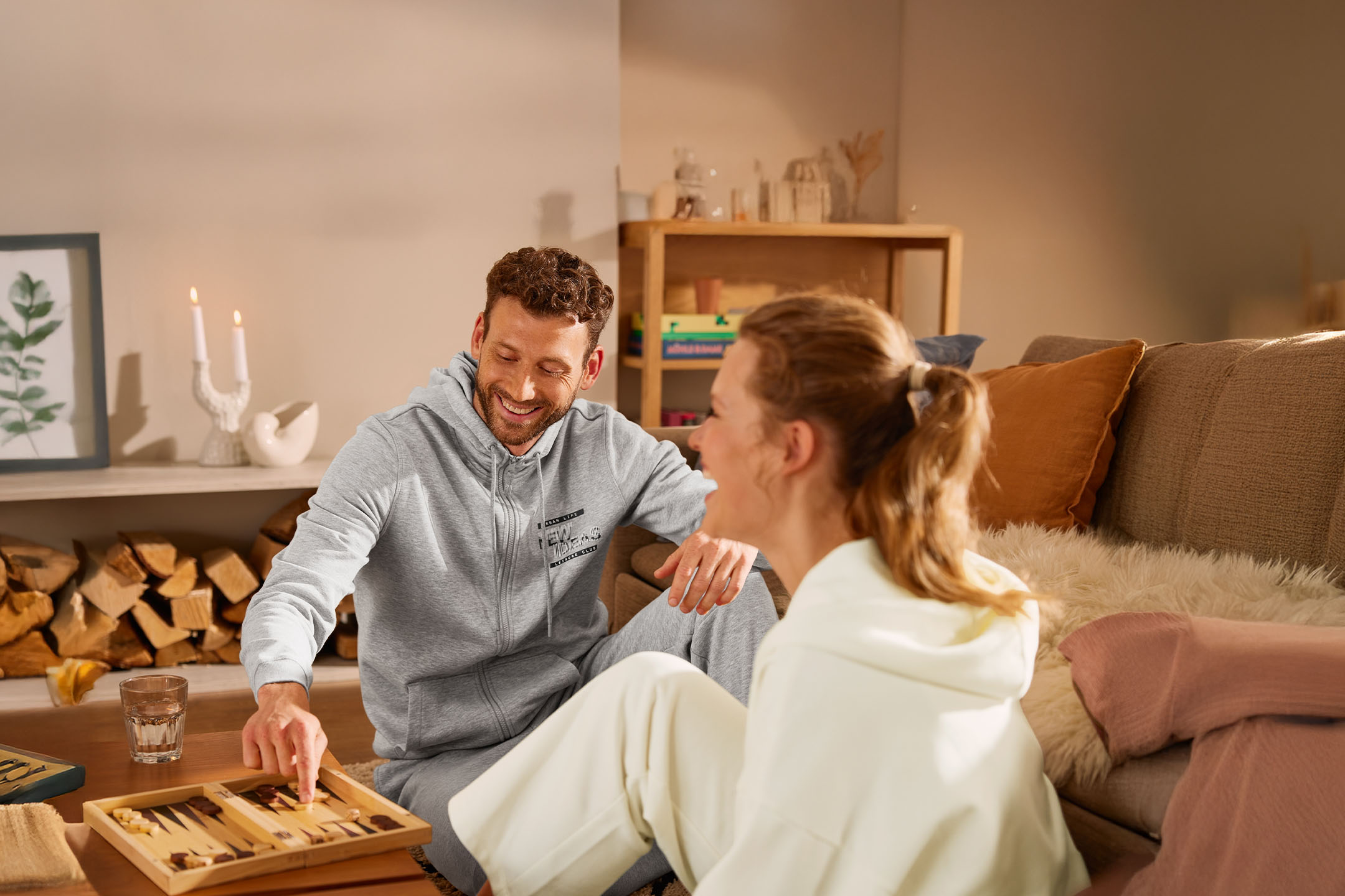 Ein Paar in Loungewear spielt Backgammon auf dem Boden, der Mann trägt einen grauen Hoodie.