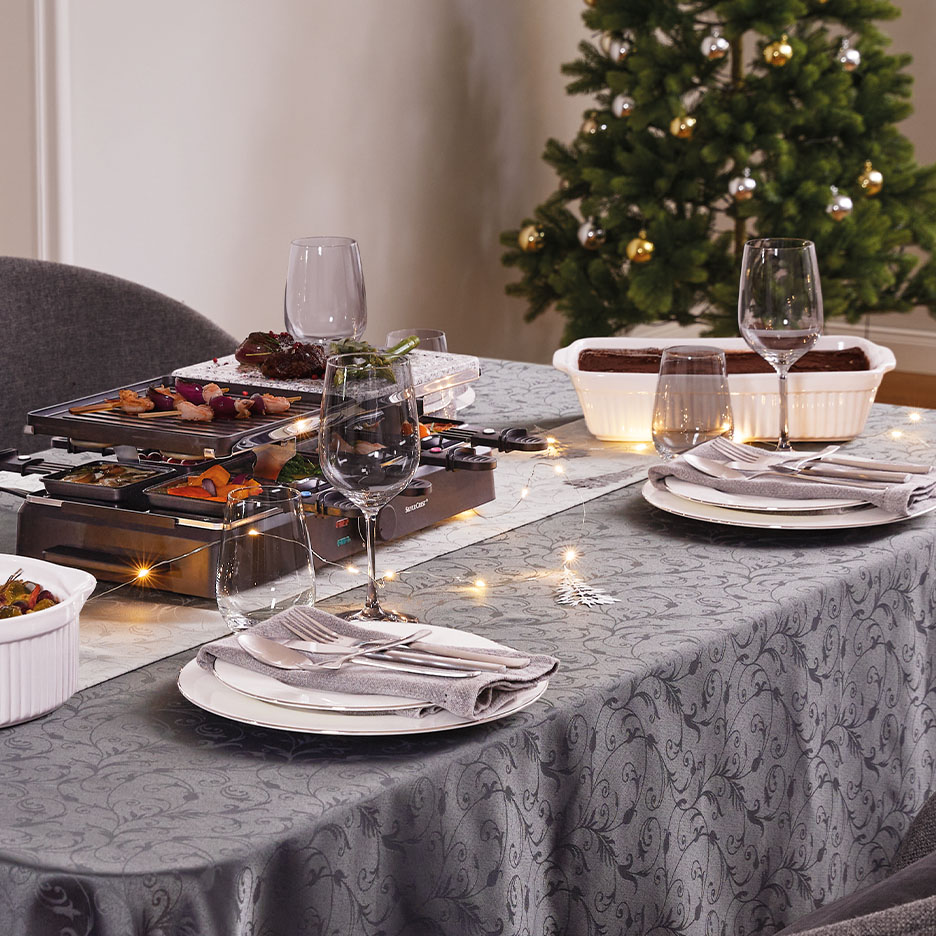 Festlich gedeckter Tisch mit Raclette-Grill, Auflaufformen und Weihnachtsbaum im Hintergrund.