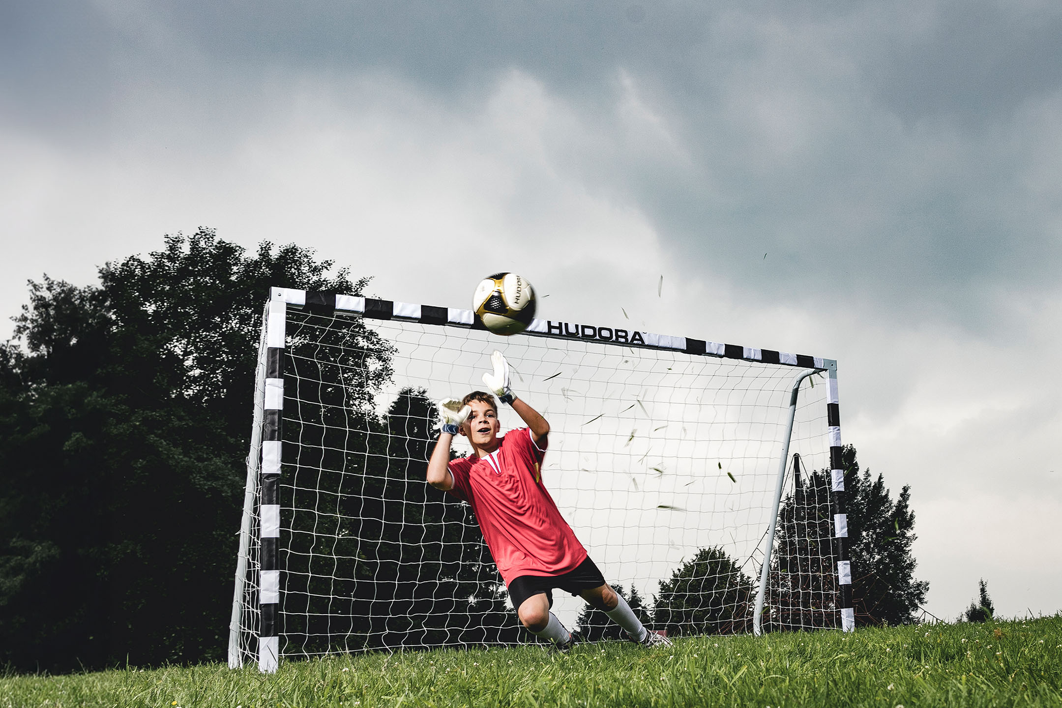 Ein junger Torwart in rotem Trikot fängt einen Fußball vor einem HUDORA Tor.