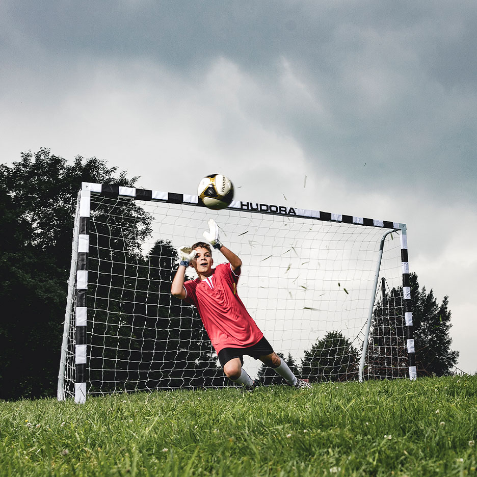 Ein junger Torwart in rotem Trikot fängt einen Fußball vor einem HUDORA Fußballtor.