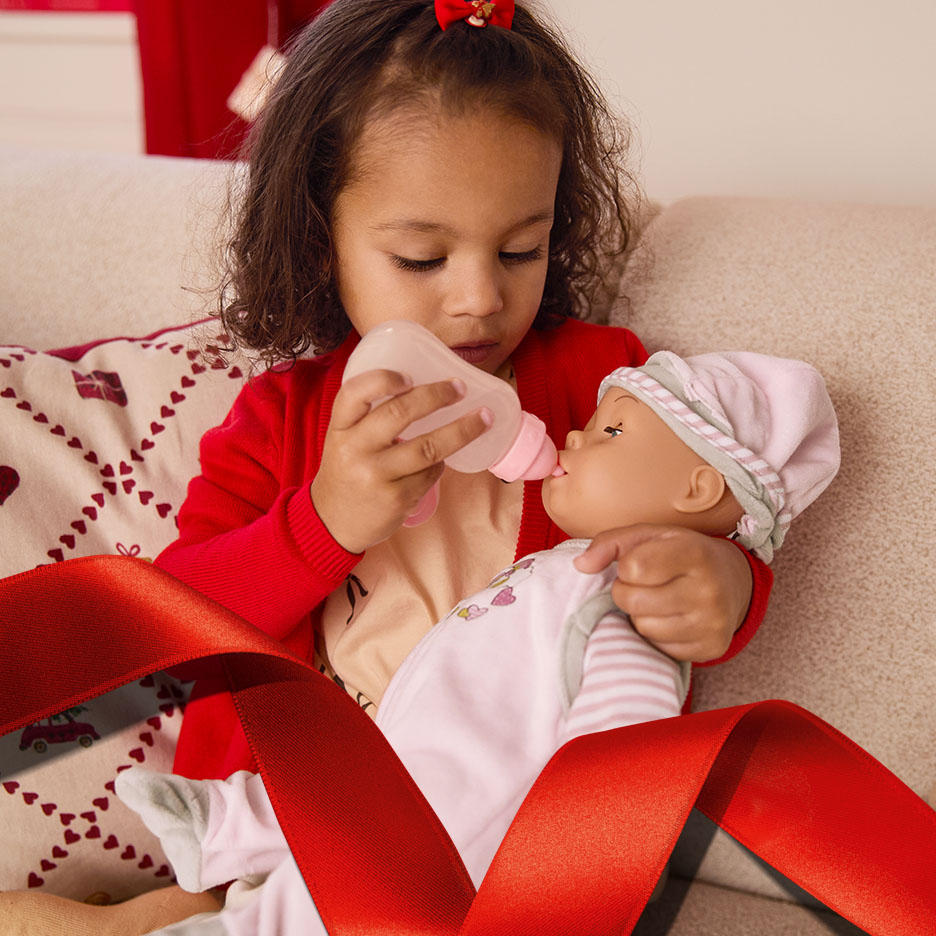 Ein kleines Mädchen füttert eine Babypuppe mit einer Flasche auf einem Sofa.