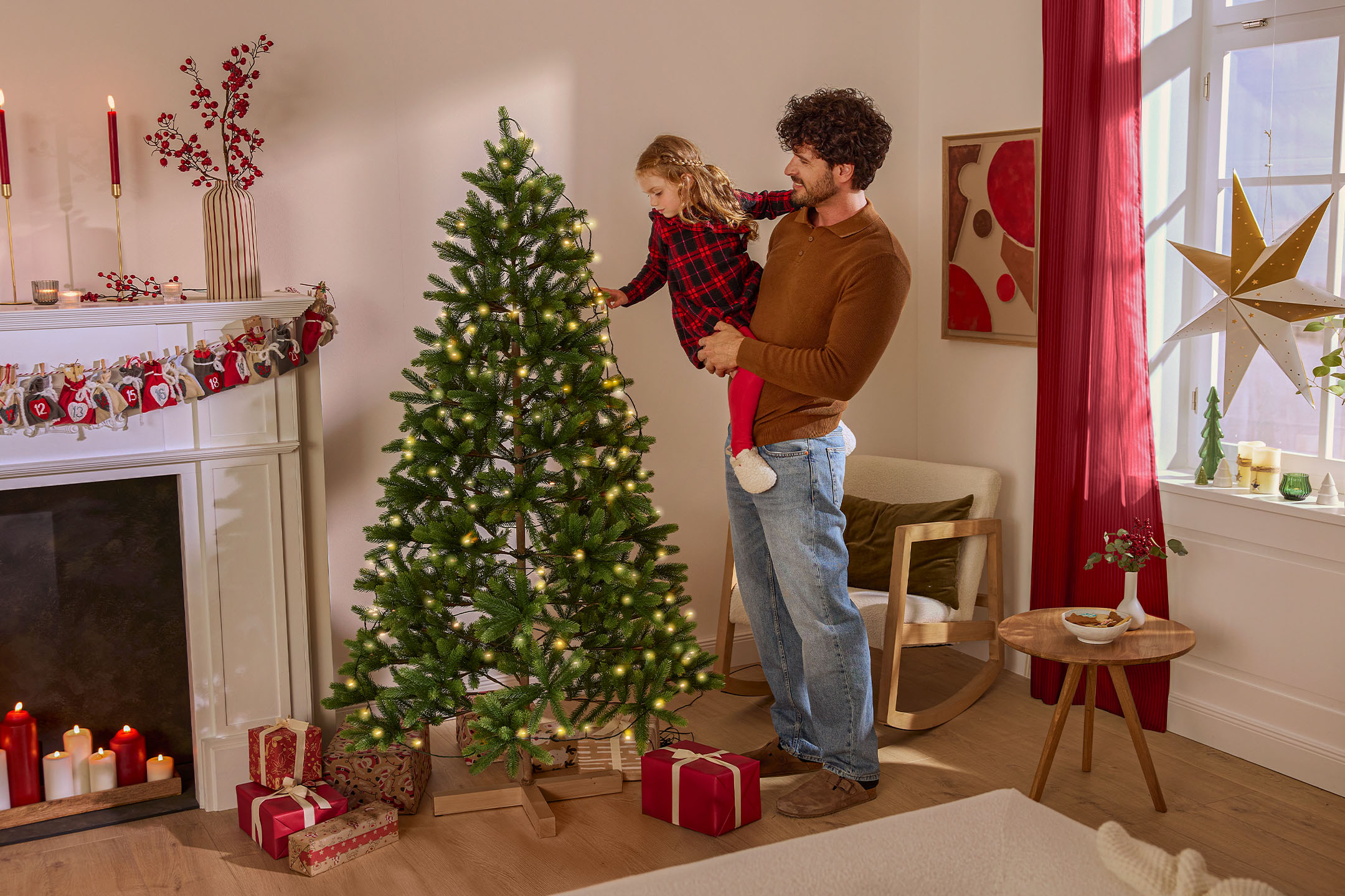 Vater und Kind schmücken einen Weihnachtsbaum mit Lichterkette, umgeben von Weihnachtsdekoration.