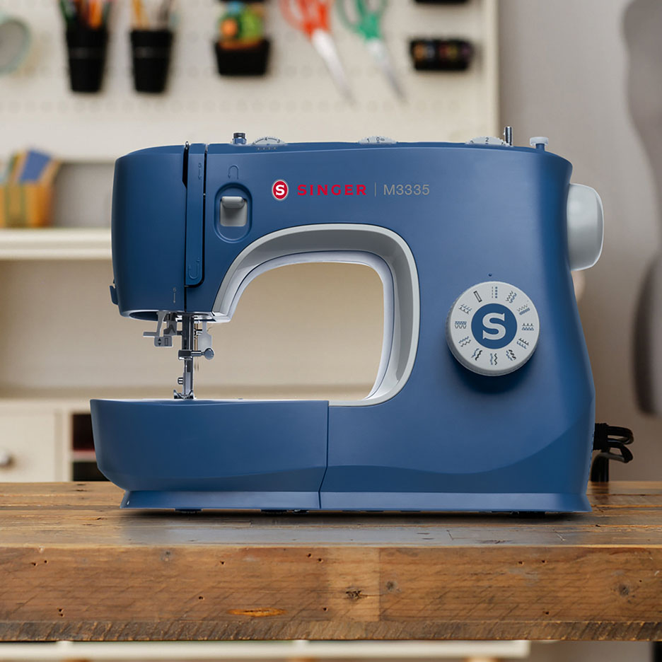 Blaue SINGER M3335 Nähmaschine auf einem Holztisch mit Bastelbedarf im Hintergrund.