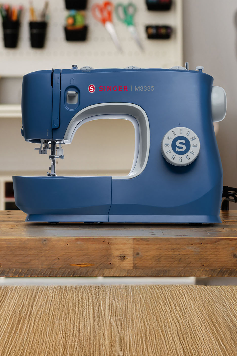 Blaue SINGER M3335 Nähmaschine auf einem Holztisch mit Bastelzubehör im Hintergrund.