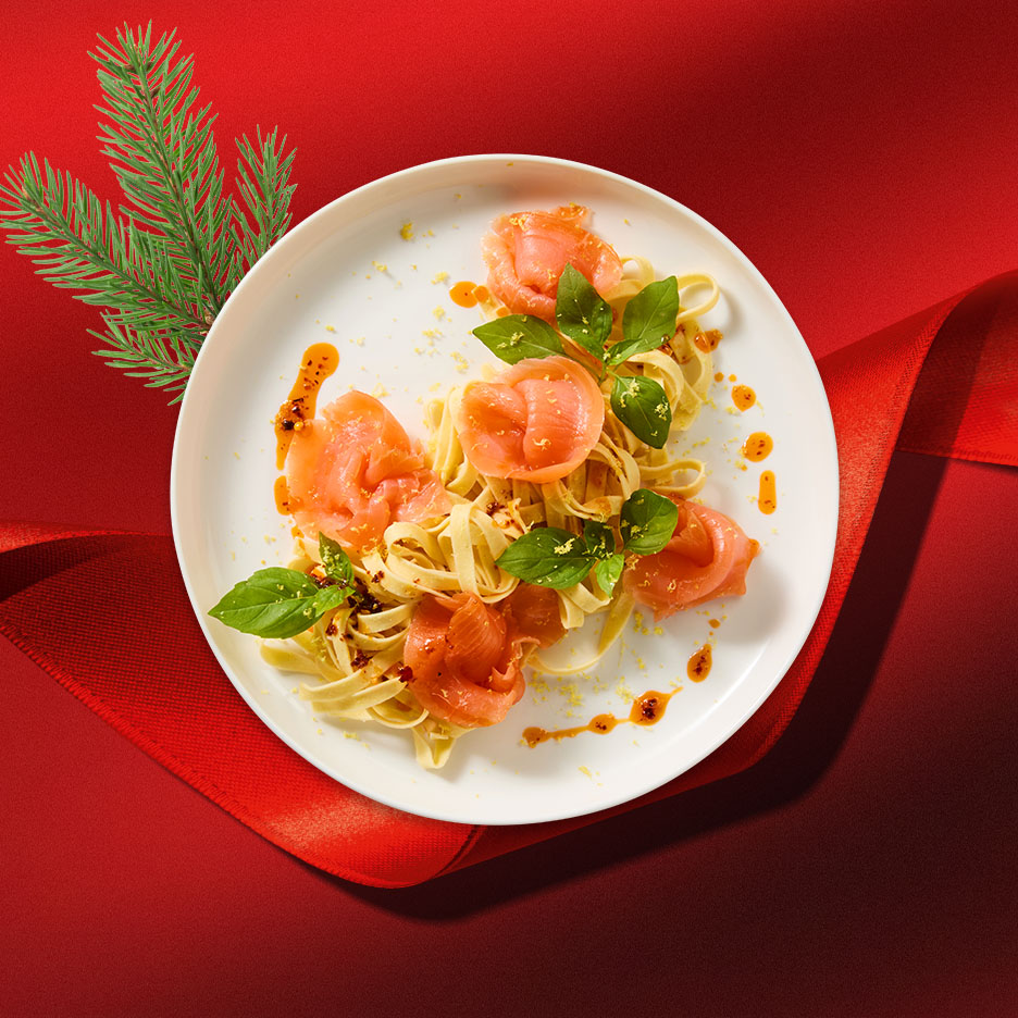 Tagliatelle mit Lachs, Basilikum und Chiliöl auf einem weißen Teller, umgeben von Weihnachtsdekoration.