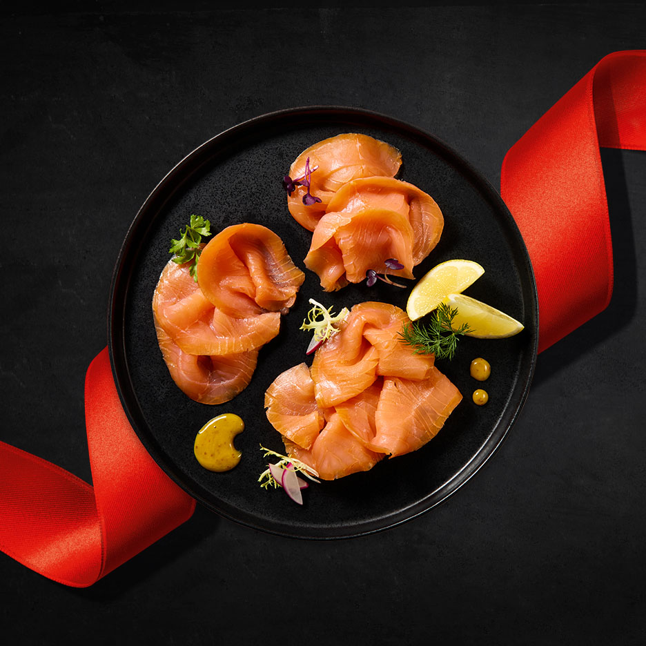 Geräucherter Lachs auf schwarzem Teller mit Zitrone und Senf, umwickelt von rotem Band.