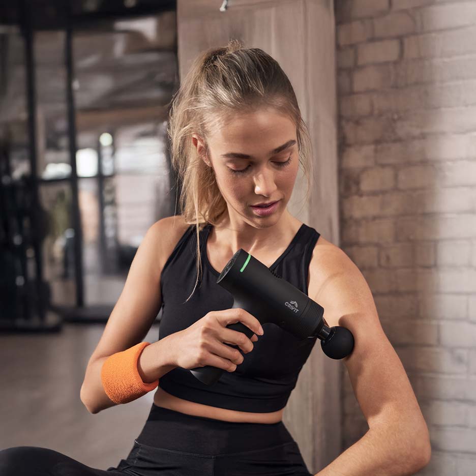 Frau massiert ihren Arm mit einem schwarzen CRIVIT Massagegerät im Fitnessstudio.