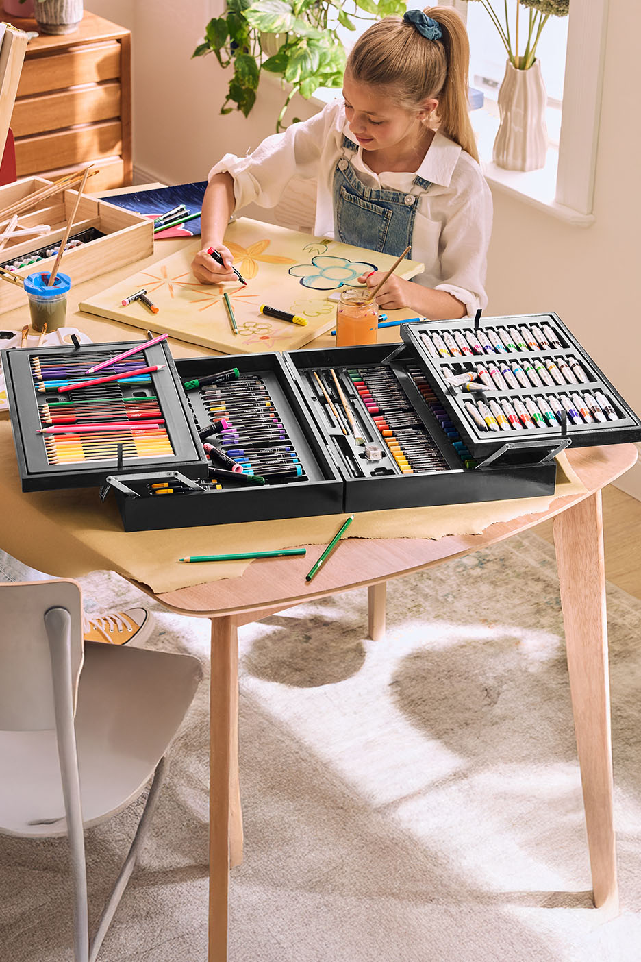 Mädchen malt mit einem großen Malset, das Buntstifte, Marker, Pinsel und Farben enthält.