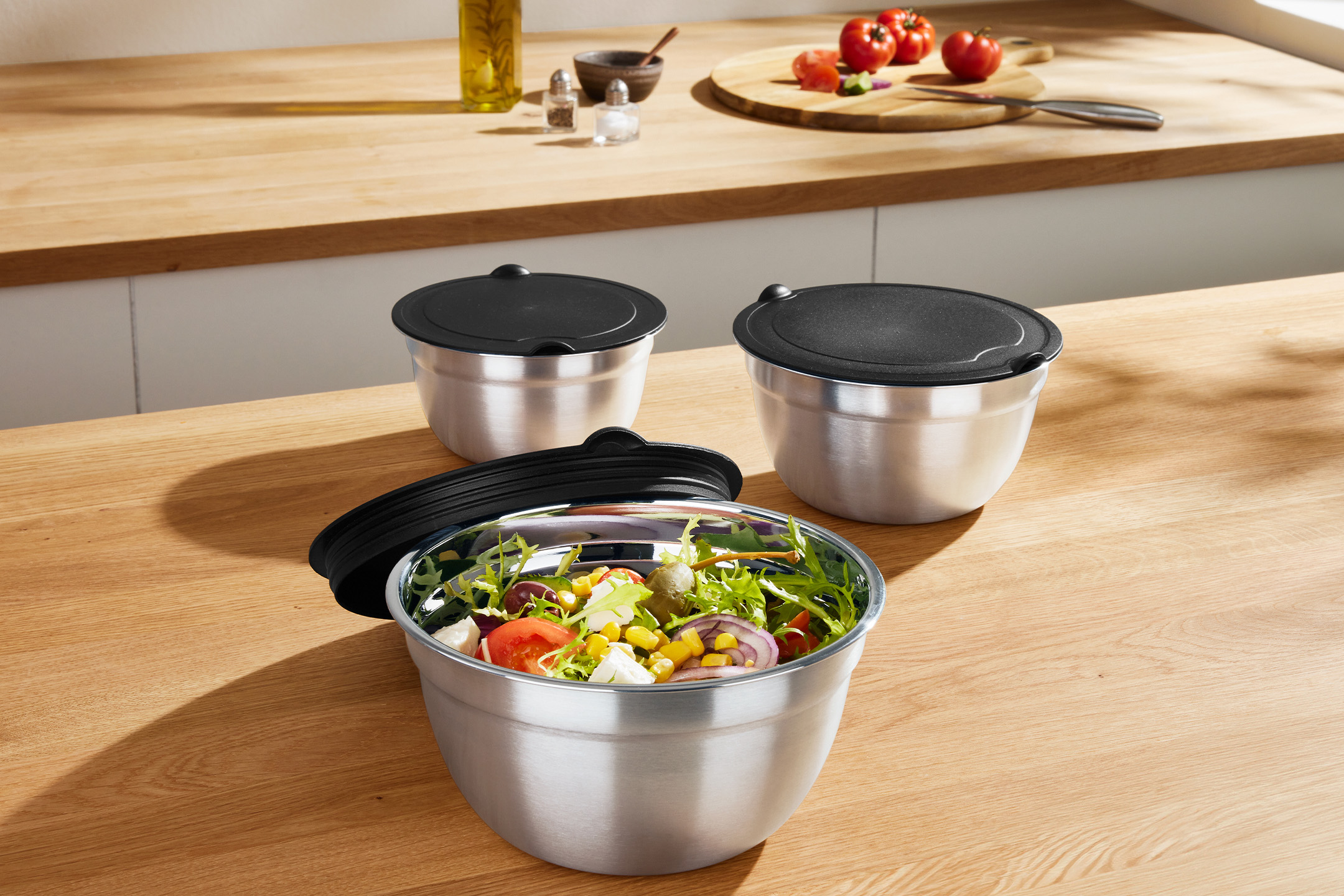 Drei silberne Salatschüsseln mit schwarzen Deckeln auf einer Holzarbeitsplatte, eine davon mit frischem Salat gefüllt.