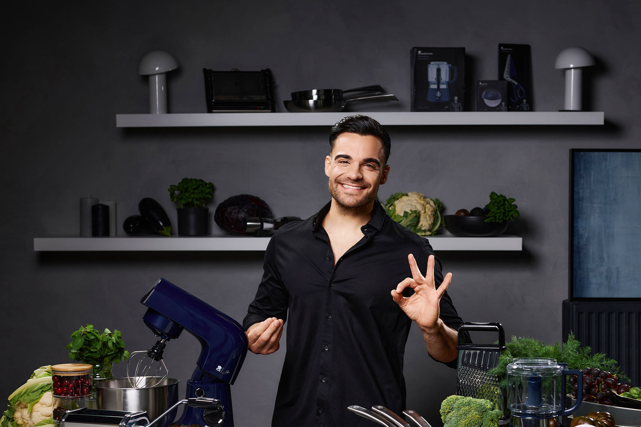 Ein lächelnder Mann in einer Küche mit einer Küchenmaschine, frischem Gemüse und Küchengeräten.