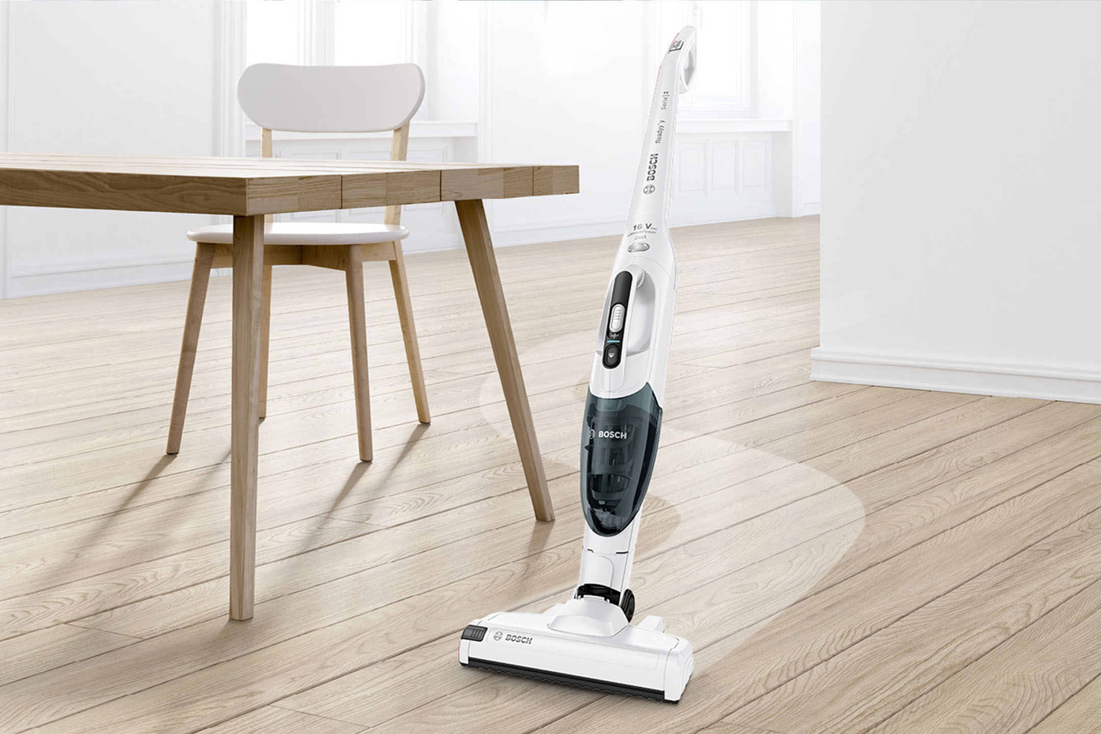 Weißer Akku-Staubsauger auf hellem Holzboden neben einem Holztisch und Stuhl