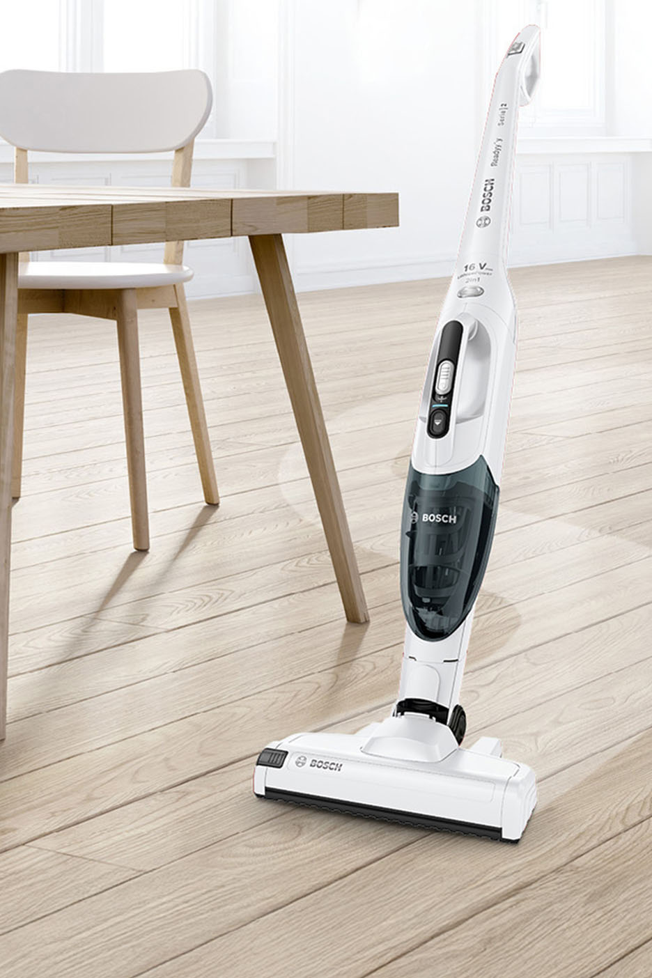 Weißer Akku-Staubsauger auf hellem Holzboden neben einem Holztisch und Stuhl.