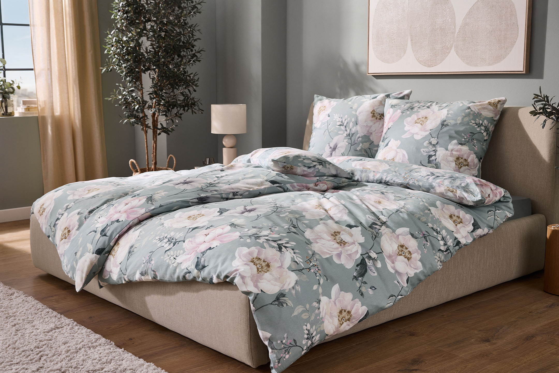 Bett mit floraler Bettwäsche in Mintgrün und Rosa, umgeben von Pflanzen und einer Lampe.