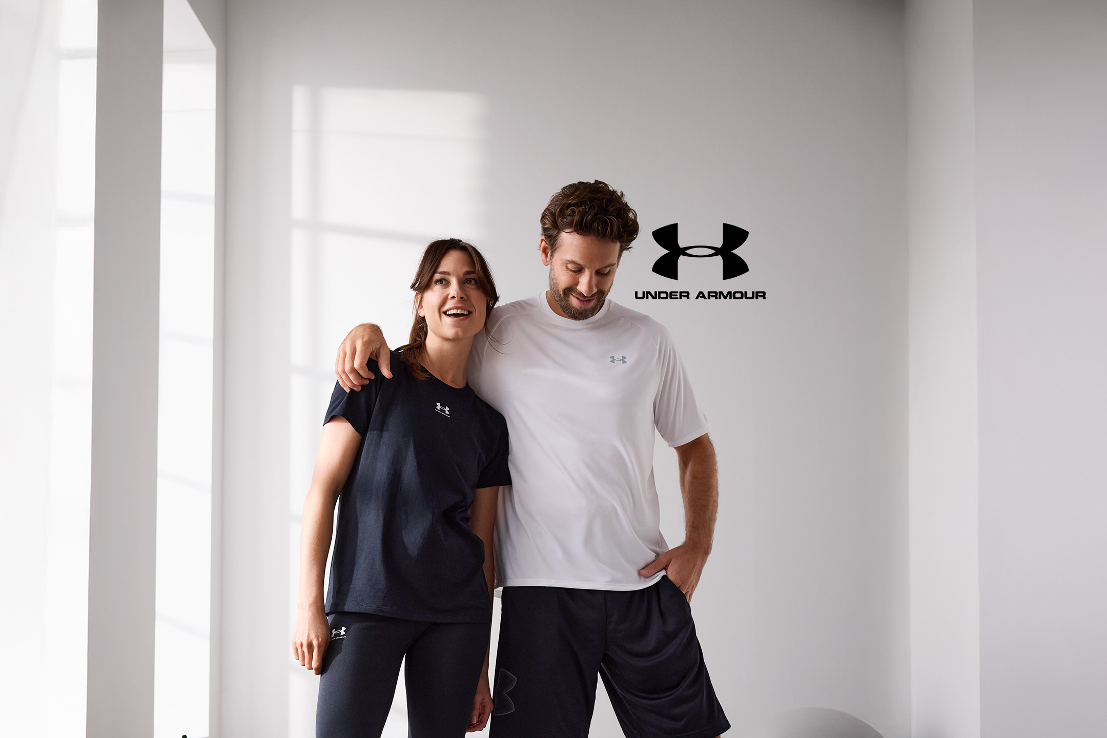 Mann und Frau in Under Armour Sportbekleidung, lächelnd vor einer weißen Wand.