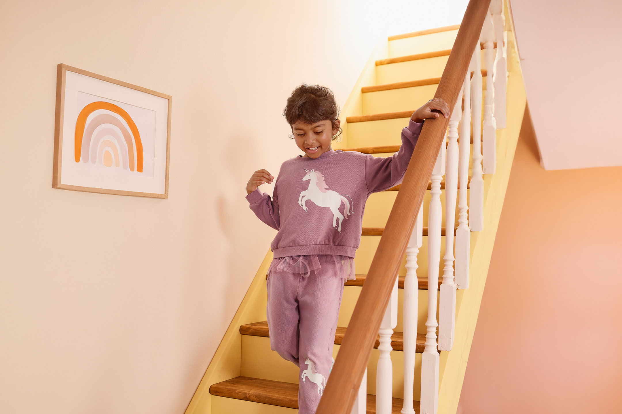 Mädchen in lila Einhorn-Pyjama auf einer Treppe mit Regenbogenbild.