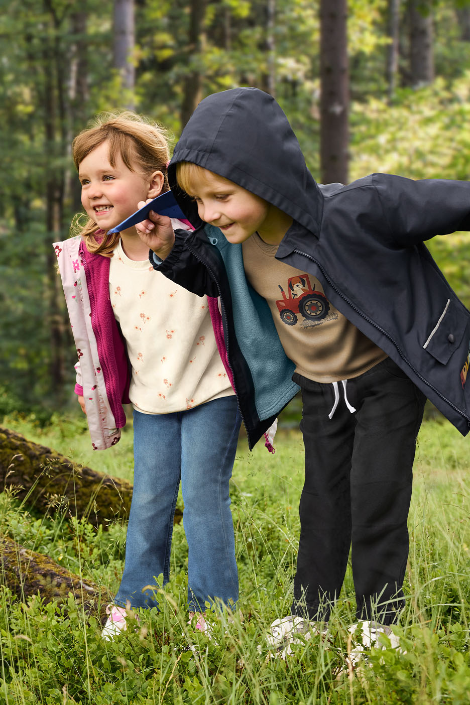 Zwei Kinder in Outdoor-Kleidung spielen im Wald, ein Junge trägt eine Kapuzenjacke und ein Mädchen eine Fleecejacke.