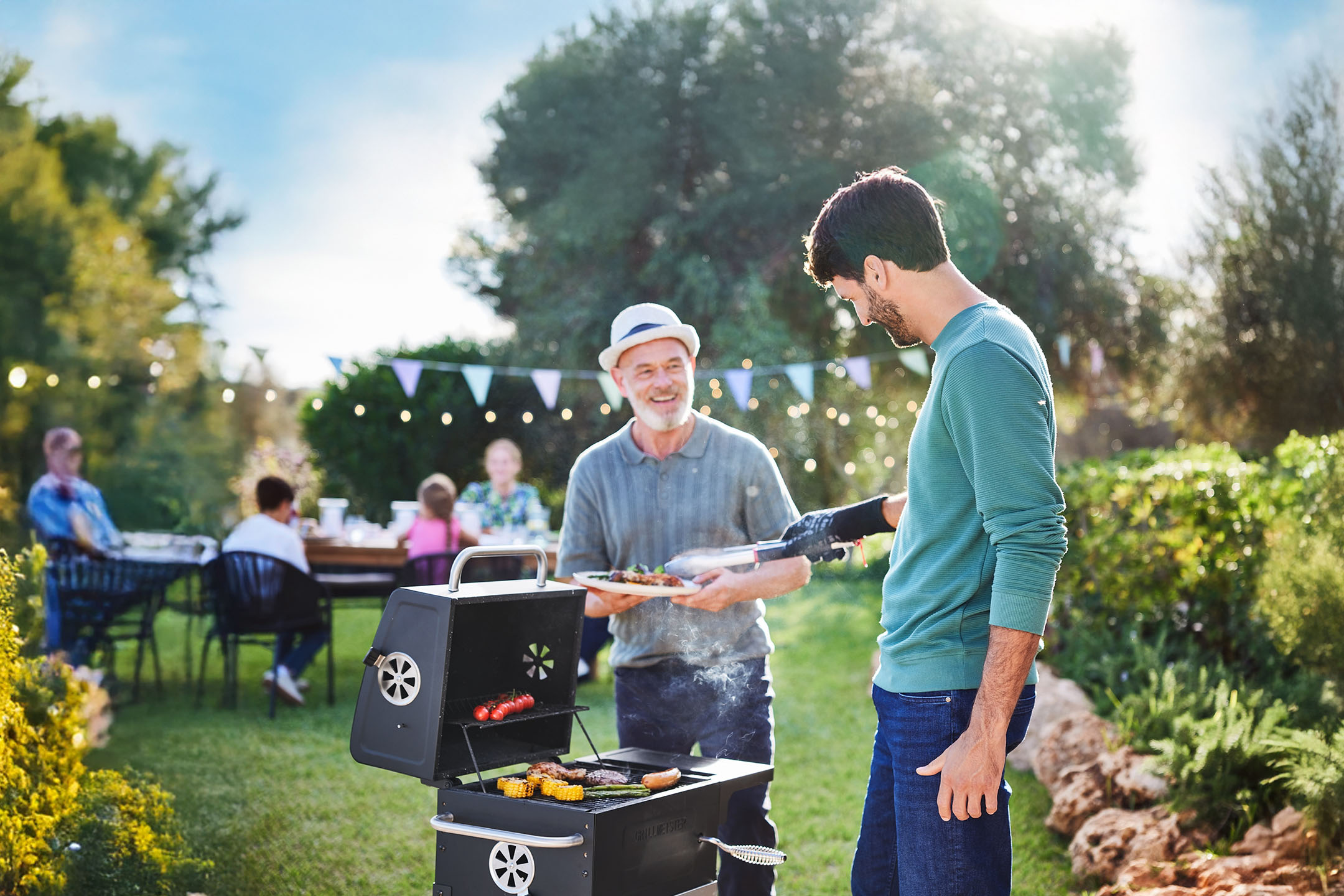 Zwei Männer grillen im Garten, während andere im Hintergrund sitzen.