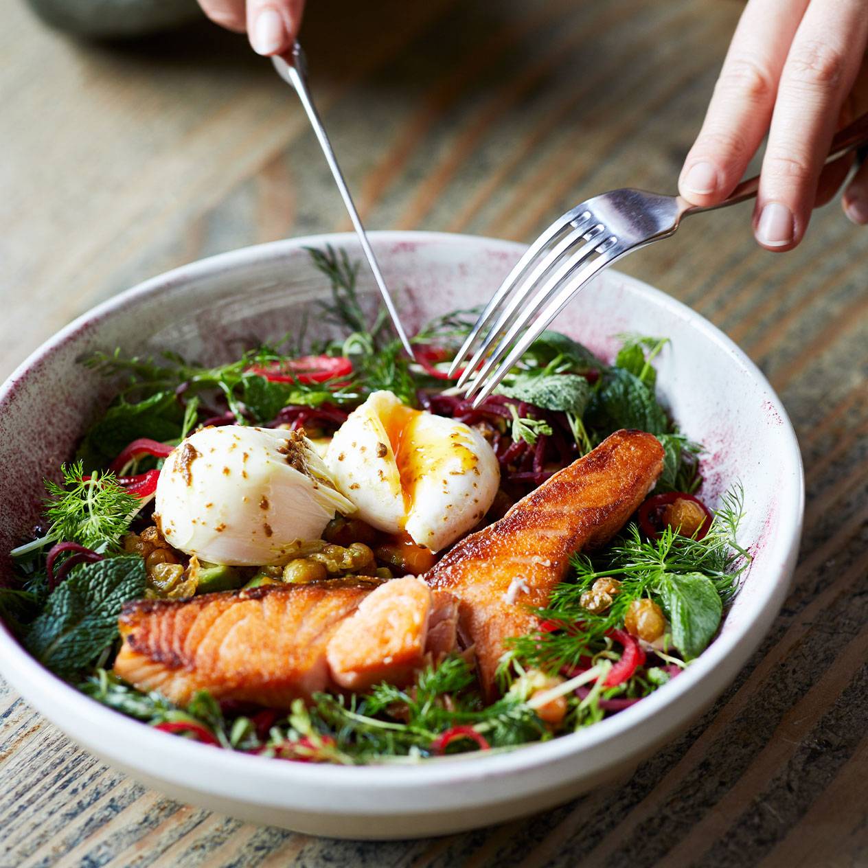 Lachs-Salat mit pochierten Eiern und frischen Kräutern in einer Schüssel, mit zwei Händen, die Messer und Gabel halten.