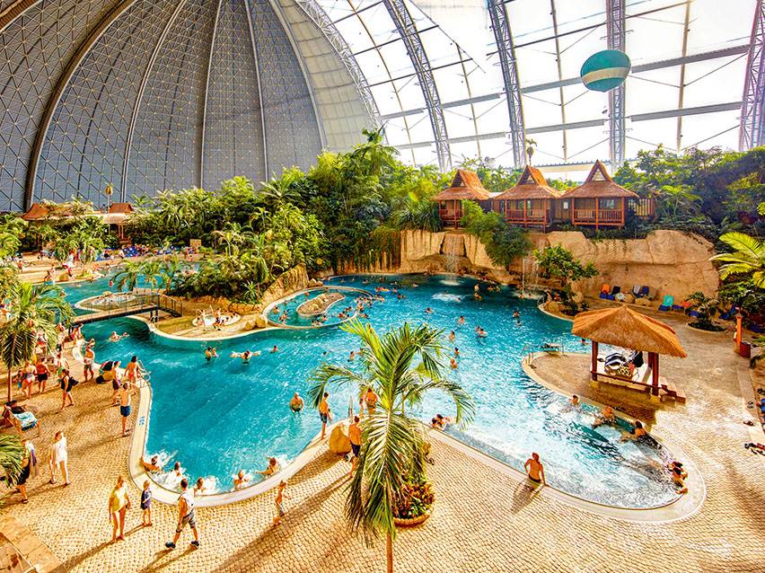 Tropische Wasserwelt mit Pools, Palmen, Hütten und vielen Menschen unter einer Glaskuppel