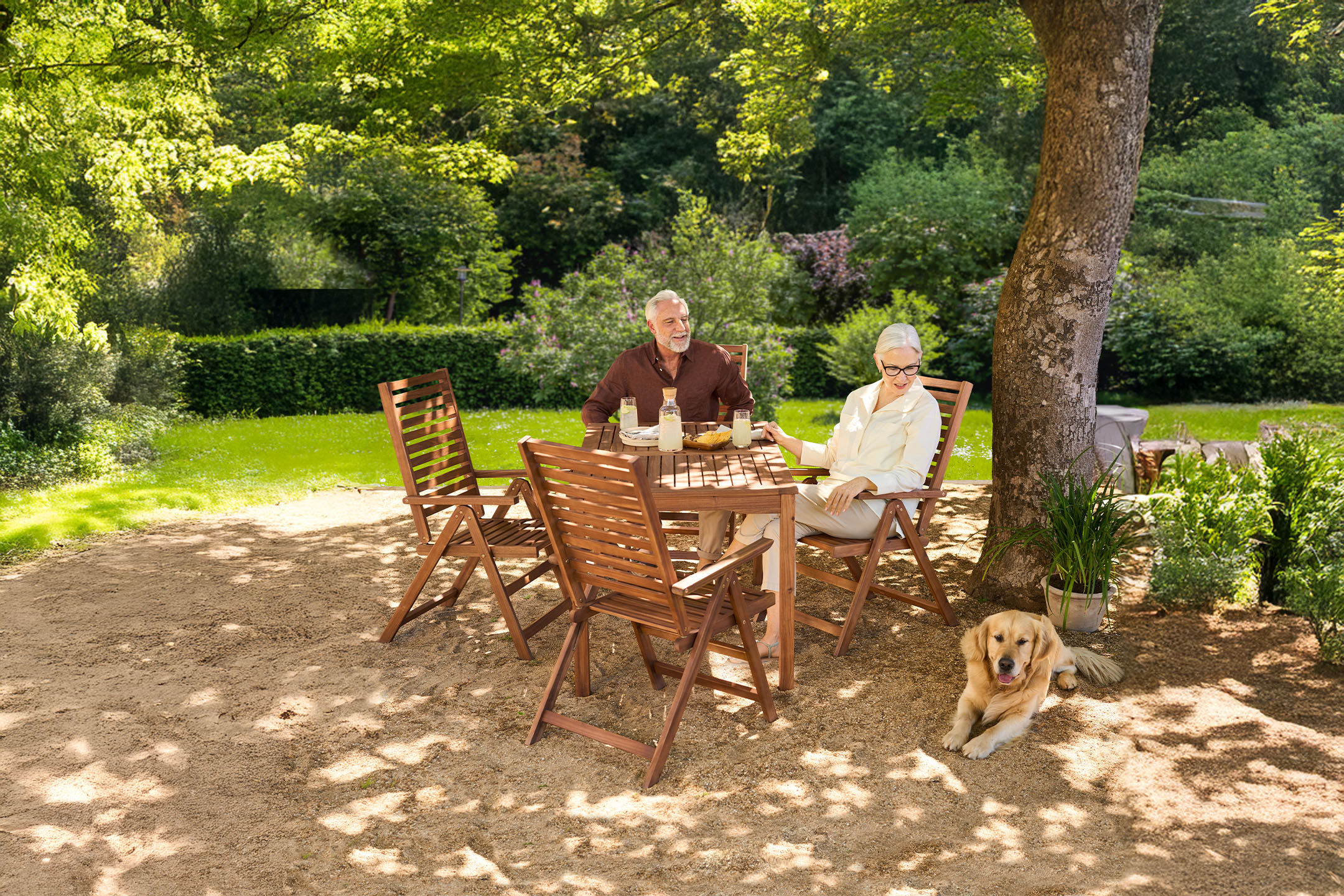 Seniorenpaar und Hund genießen ein Getränk an einem Holztisch im Garten.