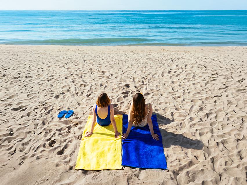 Zwei Frauen liegen auf gelben und blauen Strandtüchern am Strand und blicken auf das Meer.