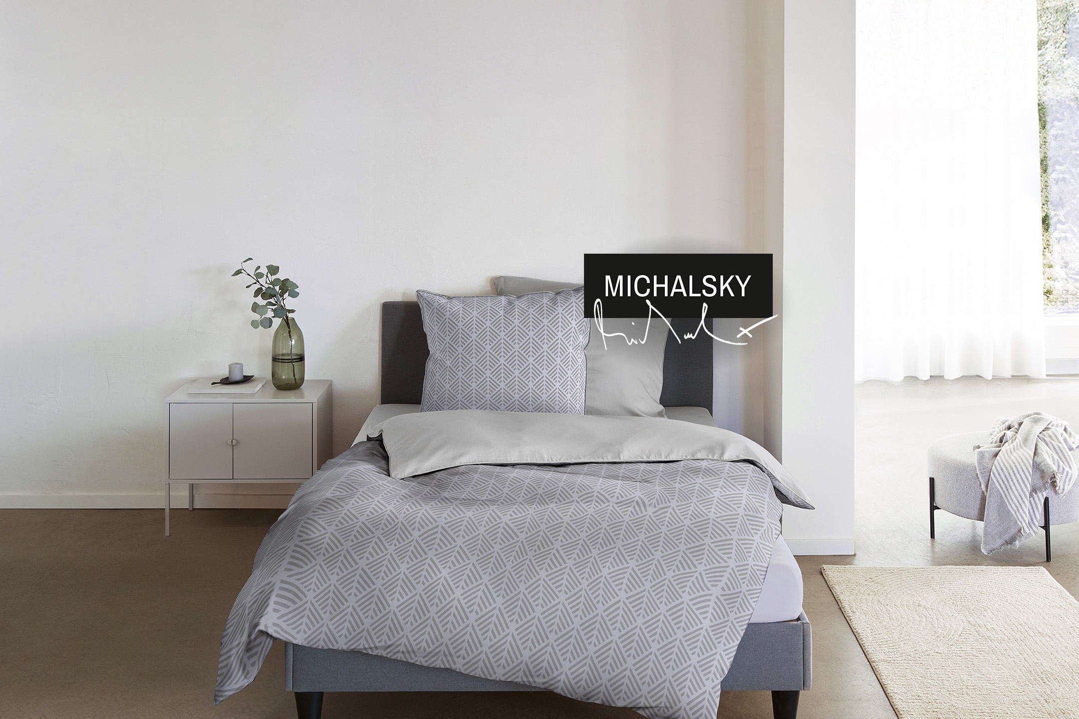 Bett mit grauer Bettwäsche und geometrischem Muster, Nachttisch mit Vase und Hocker.