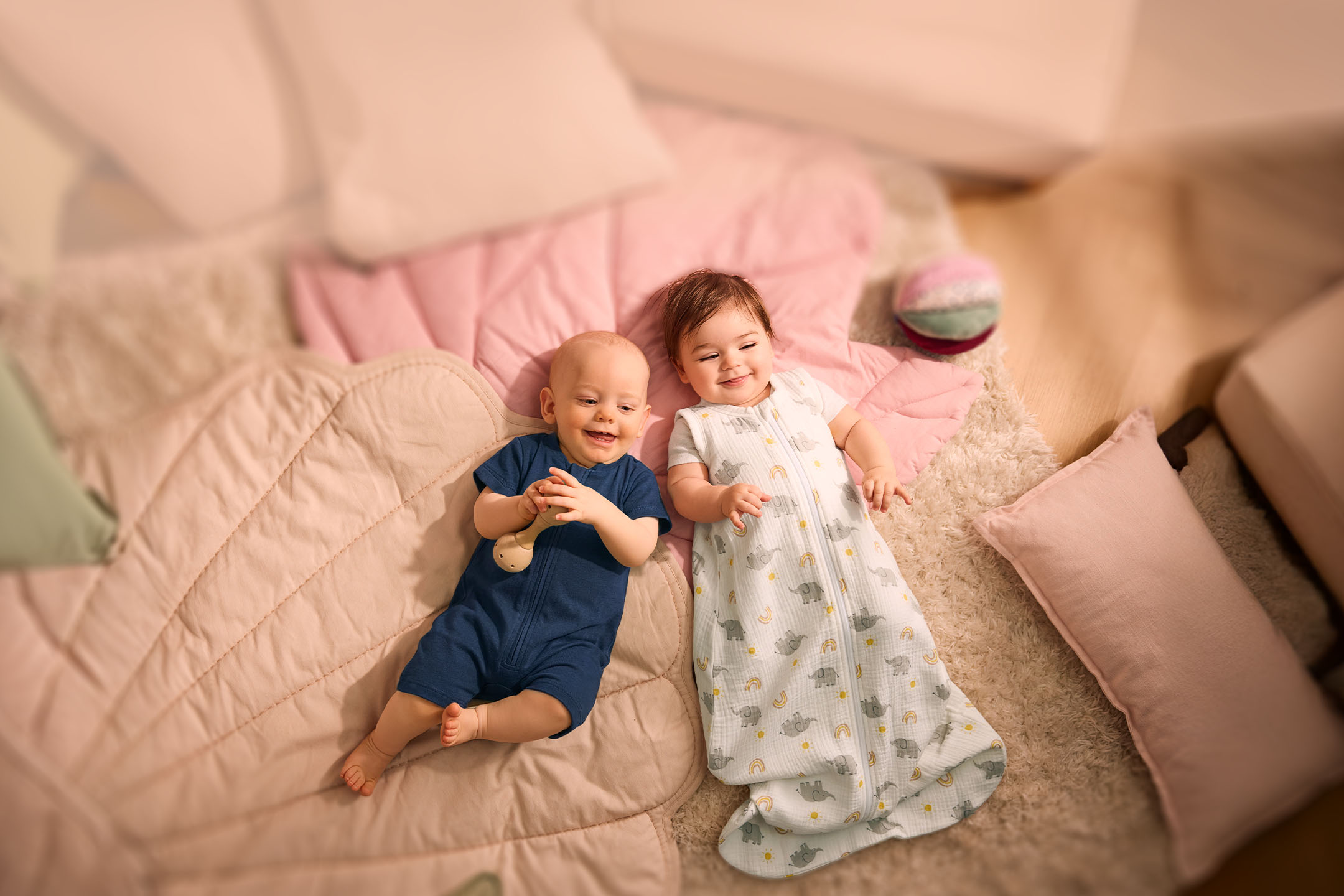 Zwei Babys liegen auf einer rosa Decke, eines in einem blauen Strampler, das andere in einem Schlafsack mit Elefantenmuster.