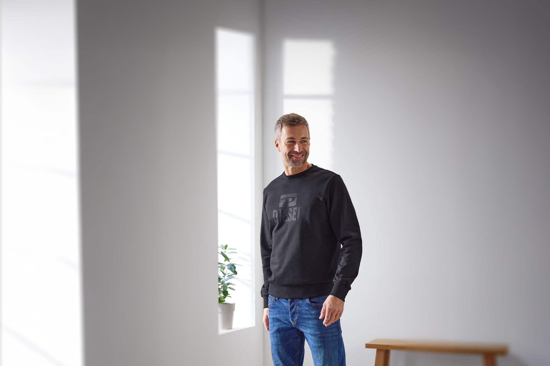 Ein lächelnder Mann in einem schwarzen Sweatshirt und Jeans steht in einem hellen Raum.