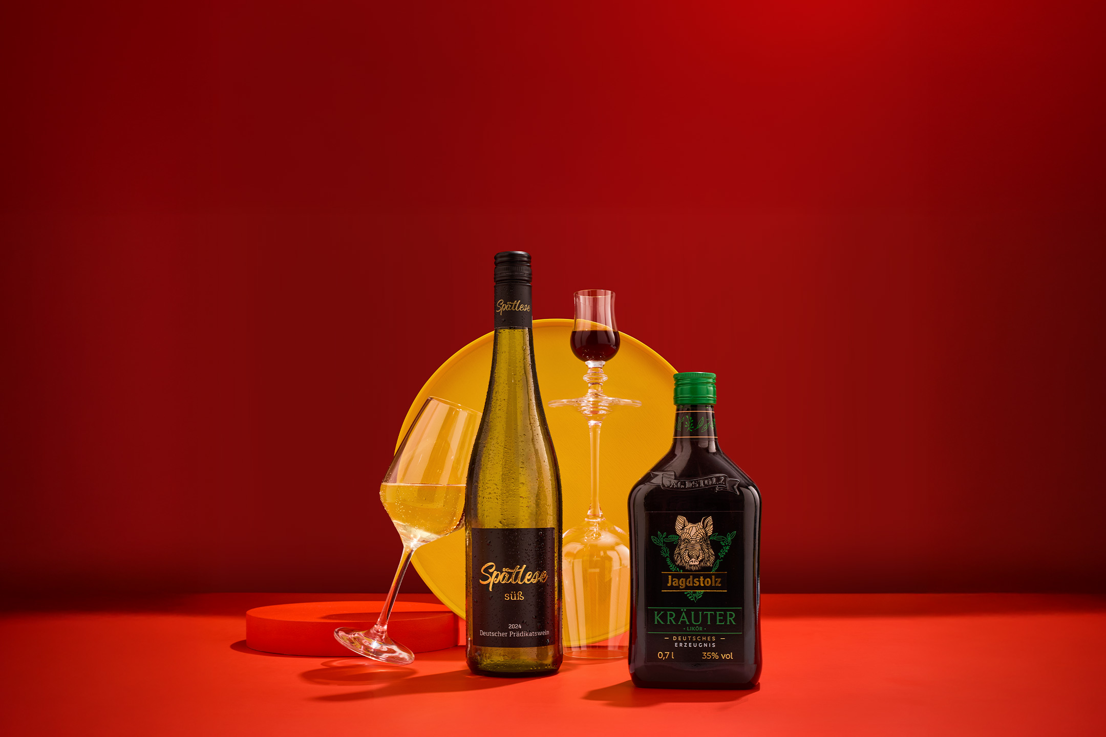 Eine Flasche Weißwein und eine Flasche Kräuterlikör mit Gläsern auf rotem Hintergrund.