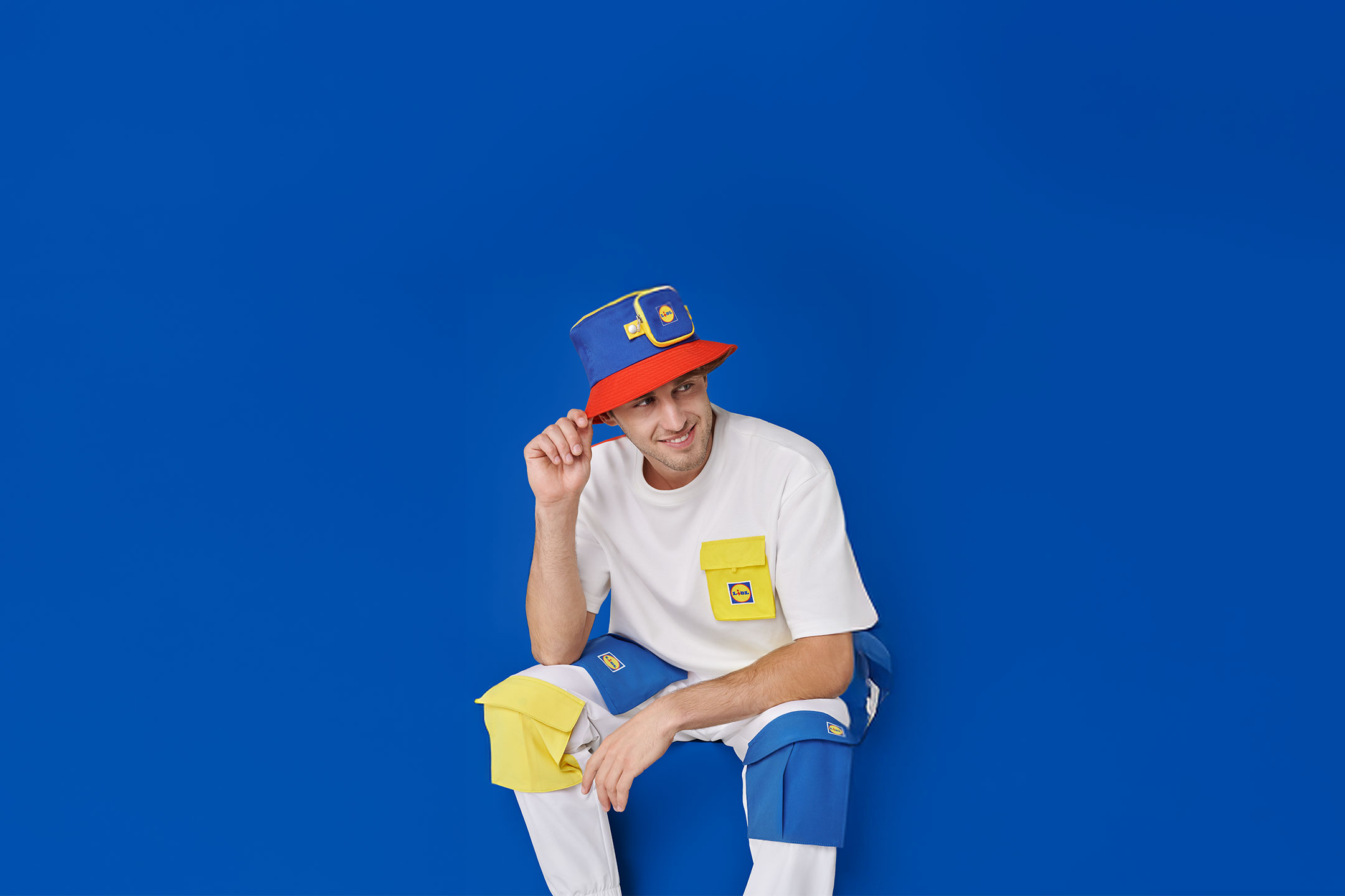 Mann in weißem T-Shirt, blauer Hose und blau-rotem Fischerhut mit gelben Taschen.