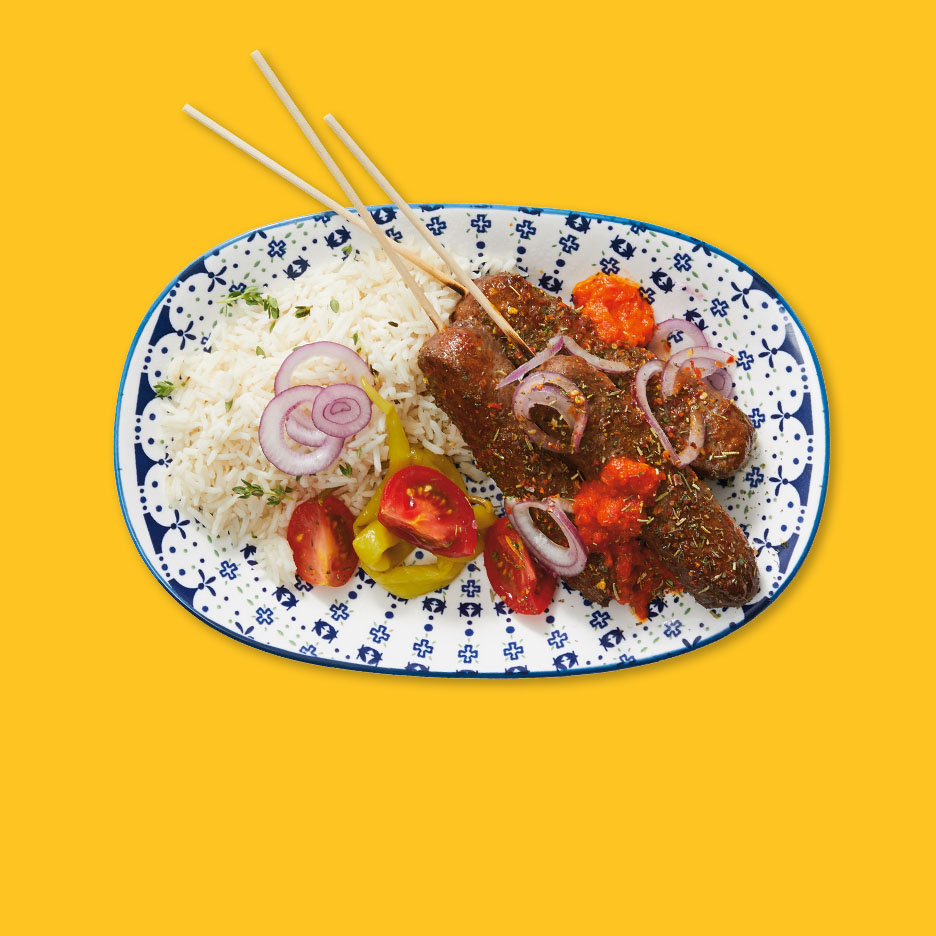 Gegrillte Köfte-Spieße mit Reis, Tomaten, Zwiebeln und Peperoni auf einem Teller.