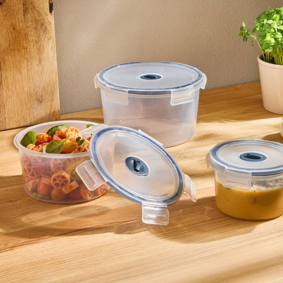 Transparente runde Lebensmittelbehälter mit blau umrandeten Deckeln, einer gefüllt mit Nudelsalat, ein anderer mit Suppe.