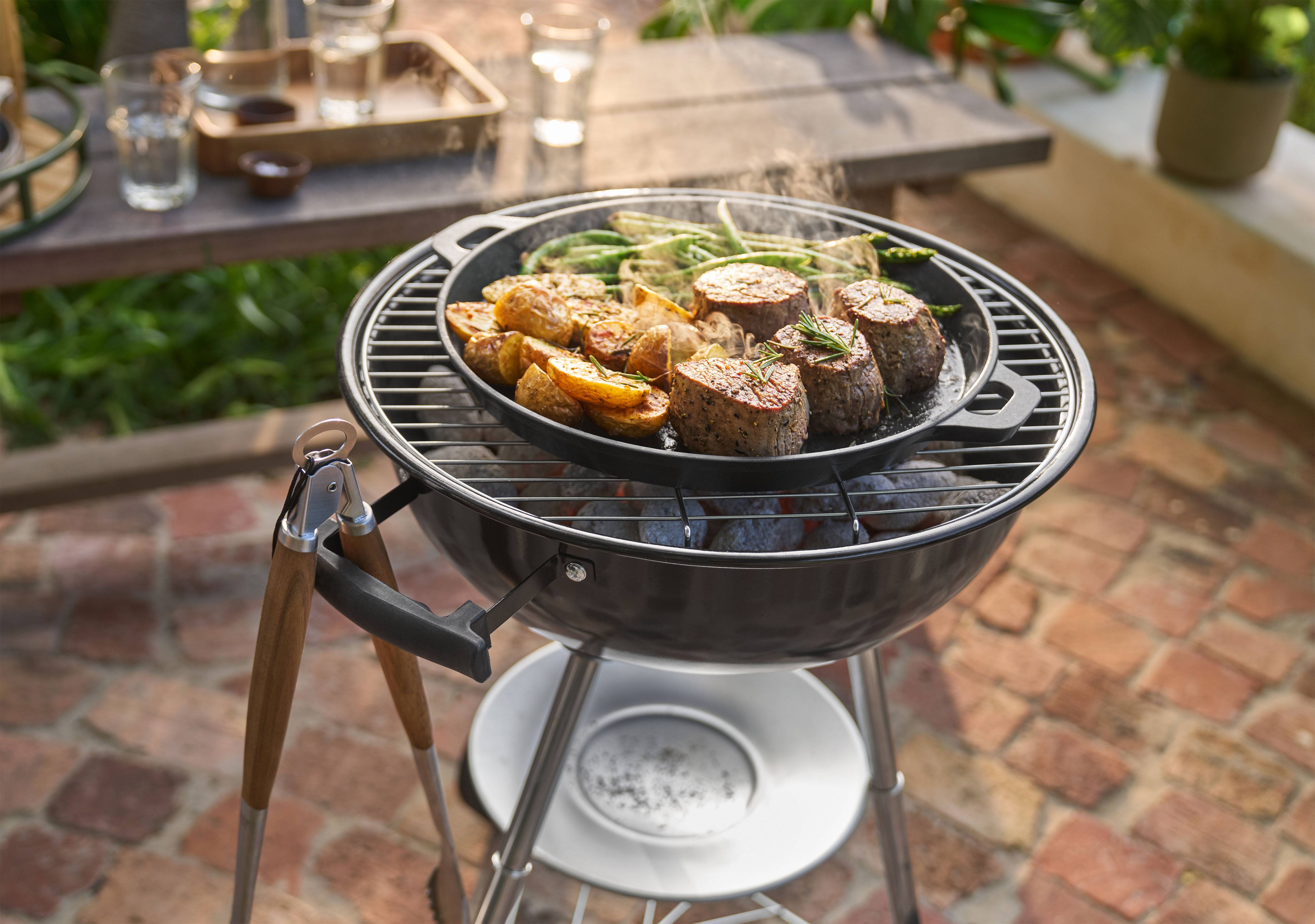 Grill mit Fleisch, Kartoffeln und grünen Bohnen, die in einer Pfanne im Freien garen.
