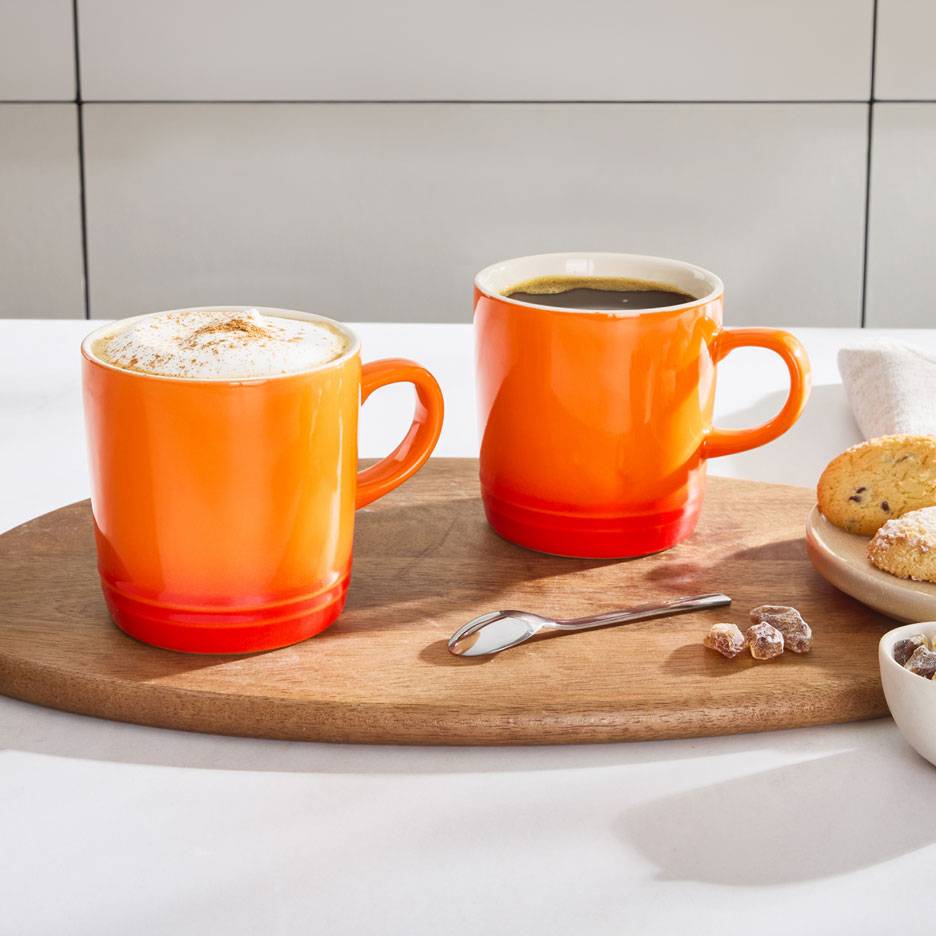 Zwei orangefarbene Kaffeetassen auf einem Holztablett, eine mit Milchschaumkaffee, die andere mit schwarzem Kaffee.