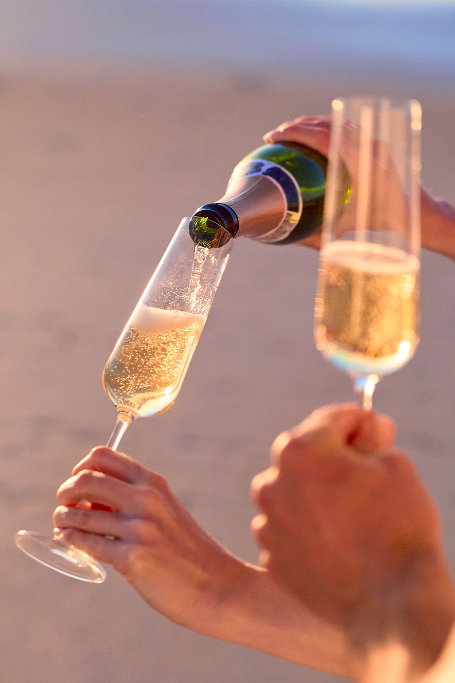 Sekt wird in zwei Gläser auf einem sandigen Hintergrund eingeschenkt.