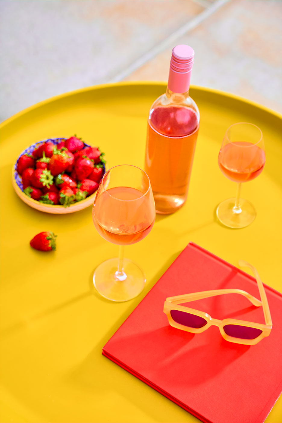 Flasche und Gläser Roséwein mit Erdbeeren und Sonnenbrille auf einem gelben Tablett.