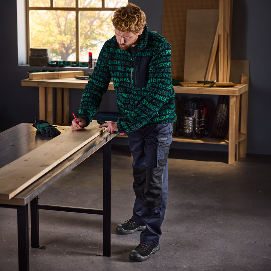 Mann in grüner Fleecejacke und Arbeitshose markiert Holz in einer Werkstatt.