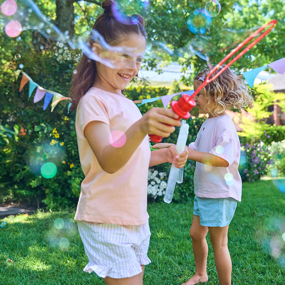Zwei Mädchen spielen mit Seifenblasenstäben in einem Garten mit bunten Wimpeln.