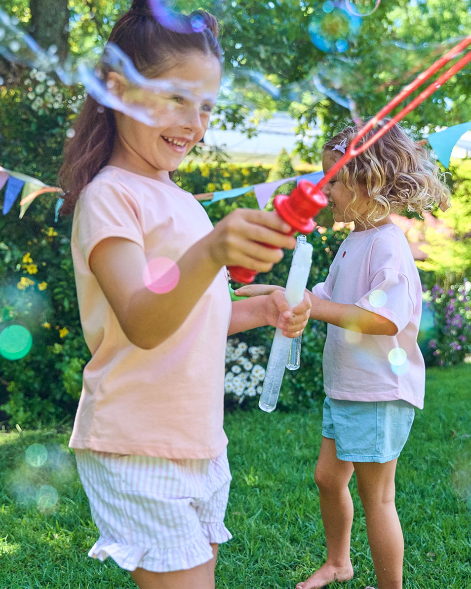 Zwei Mädchen spielen mit Seifenblasen im Garten, in Sommerkleidung.