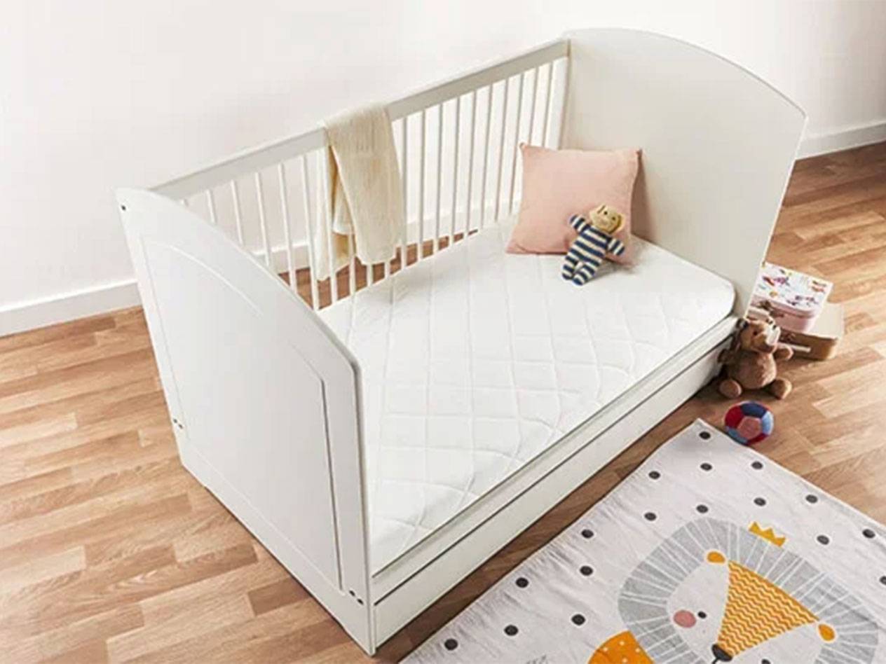 Weißes Babybett mit Matratze, Kissen und Spielzeug auf Holzboden.