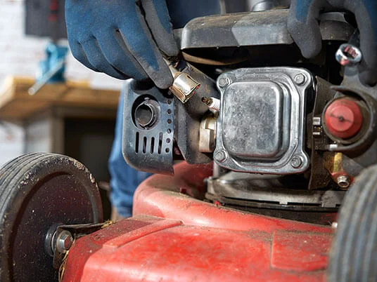 Behandschuhte Hände wechseln die Zündkerze eines roten Rasenmähers.