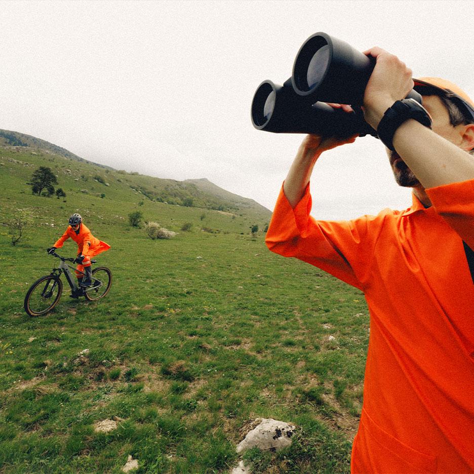 Mann mit Fernglas und Radfahrer in orangefarbener Sportkleidung in Berglandschaft.