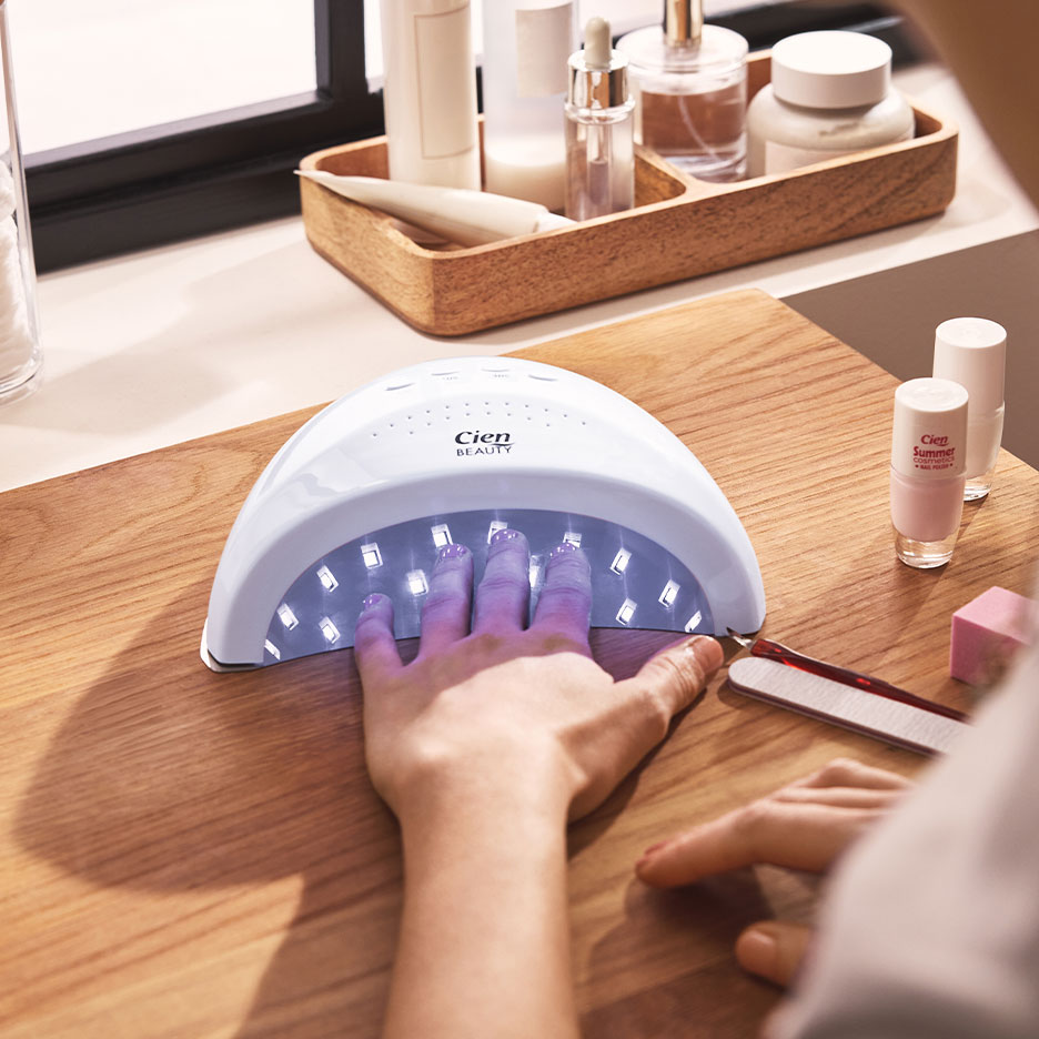 Frau trocknet Nägel in UV-Lampe, daneben Nagellacke und Maniküre-Zubehör.